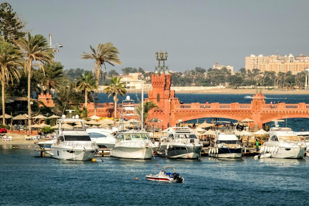 Boats moored in Port Said harbor with a red brick fortress and sandy beach lined with palm trees in the background.
