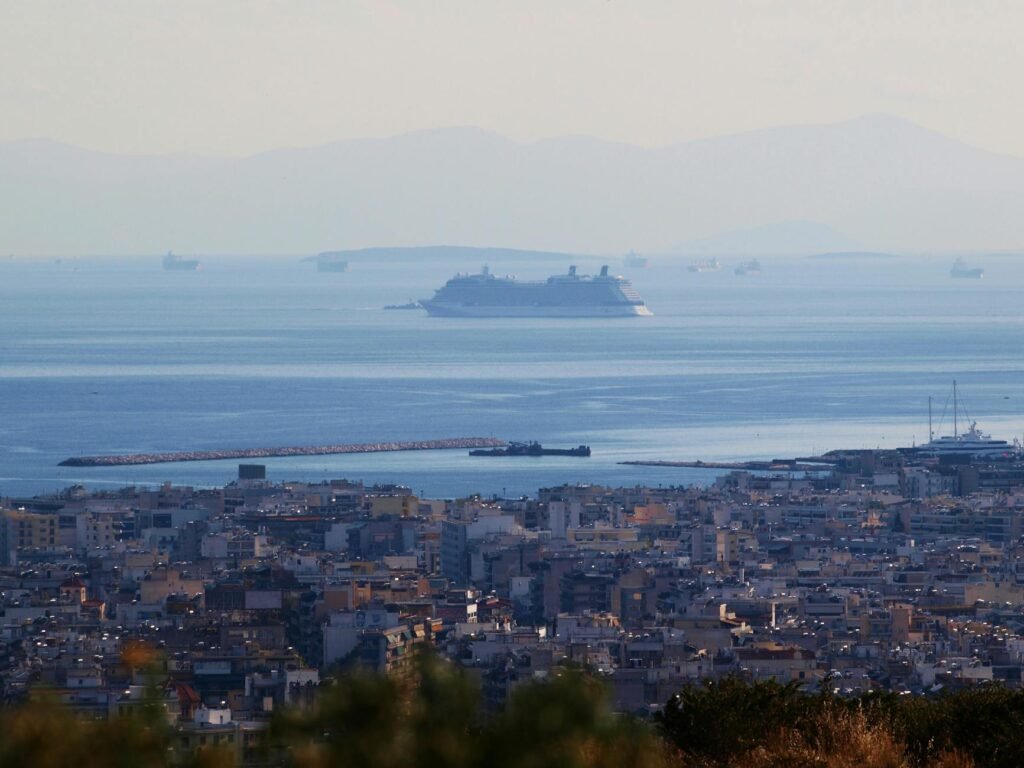 Large cruise ship anchored in Piraeus harbor with coastal city and mountains visible across the blue sea.