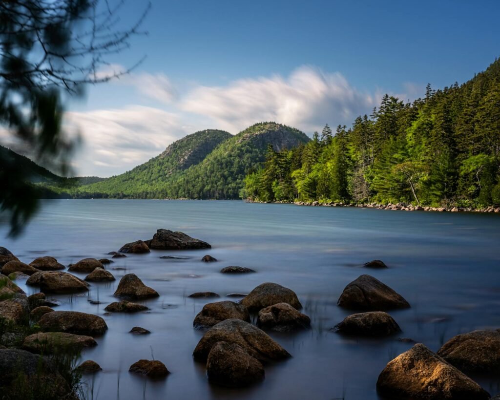 Rocky shoreline with boulders in calm lake surrounded by forested mountains under blue sky.