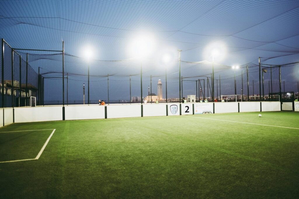 Indoor soccer field with artificial grass, netting, and city skyline visible beyond the enclosed facility.