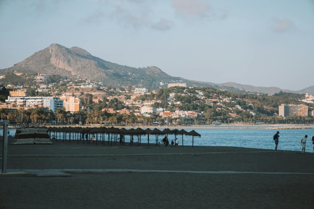 Málaga beach with black sand, beachfront pier, and mountain backdrop with hillside buildings and palm trees.