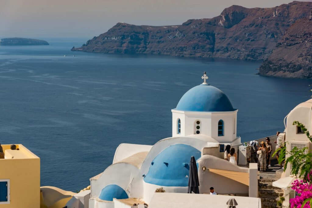 White-domed church with blue cupolas overlooking the Aegean Sea and volcanic cliffs in Santorini, Greece.
