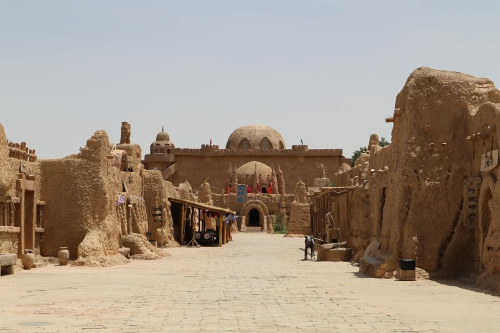 Ancient mud brick ruins of a desert town with archways and a mosque dome visible in the background.