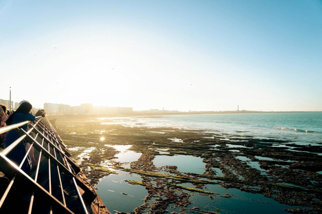 Person leaning on railing overlooking tidal pools and sandy beach in Casablanca at sunset.