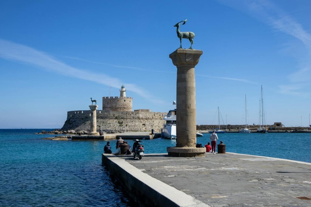 Bronze deer statue on pier with ancient fortress and sailboats in turquoise harbor at Rhodes.