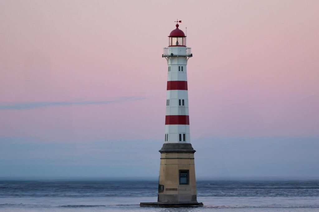 Red and white striped lighthouse standing in shallow coastal waters at dusk with pink sky.