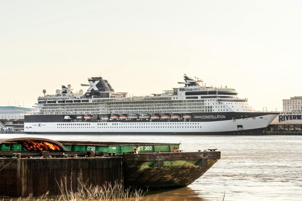 Large cruise ship docked at port with green cargo barge in foreground and brown water.