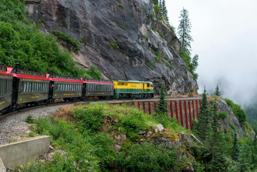 Scenic train traveling along a narrow mountain rail carved into a steep rocky cliff with dense forest and mist.