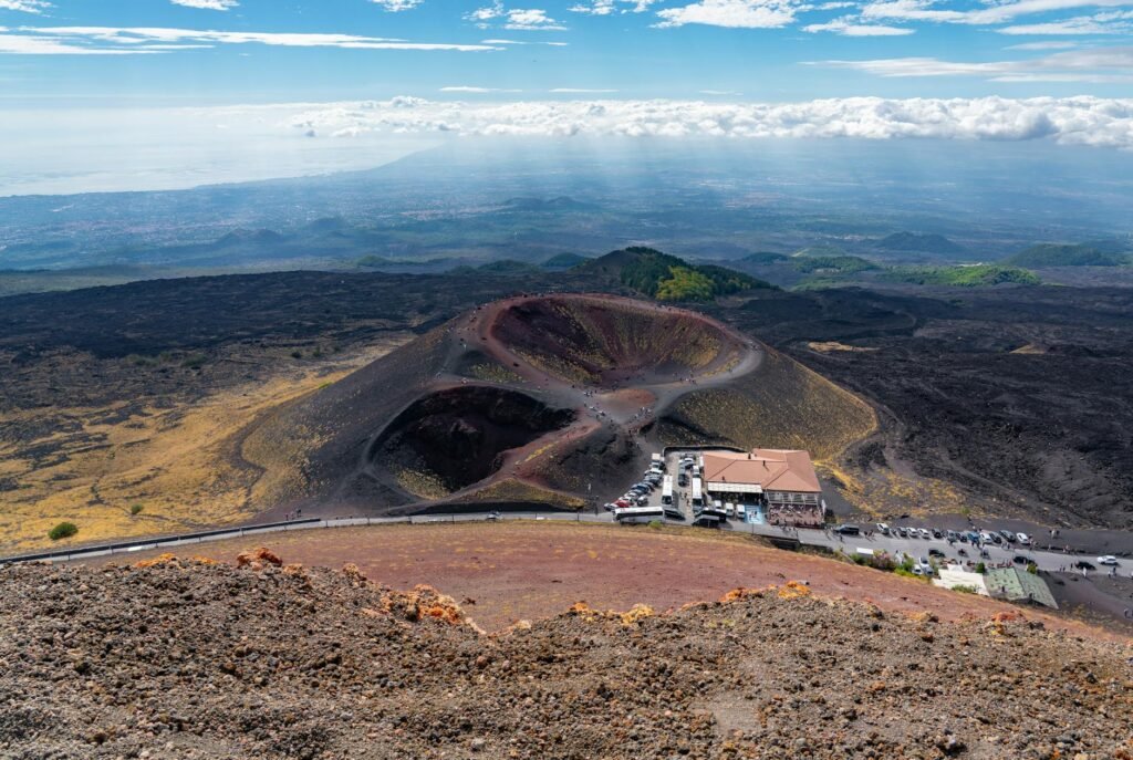 Aerial view of Mount Etna's volcanic crater with dark lava fields, visitor center, and distant Sicilian landscape.