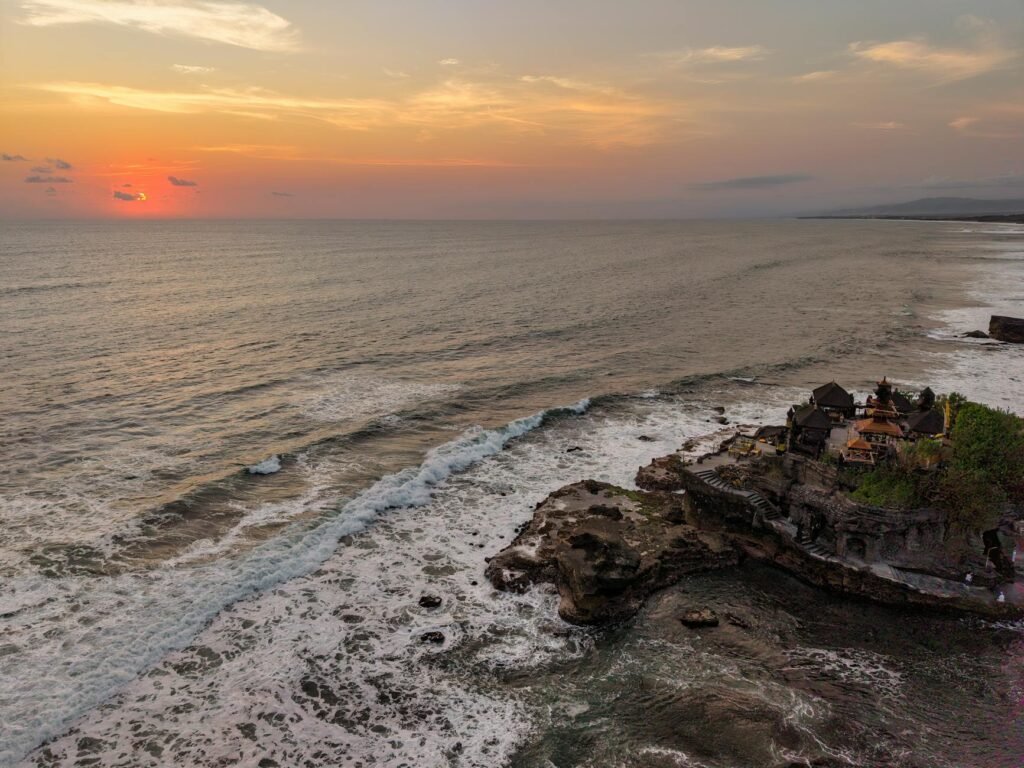 Sunset over ocean waves with rocky cliffside temple and distant coastline in Bali.