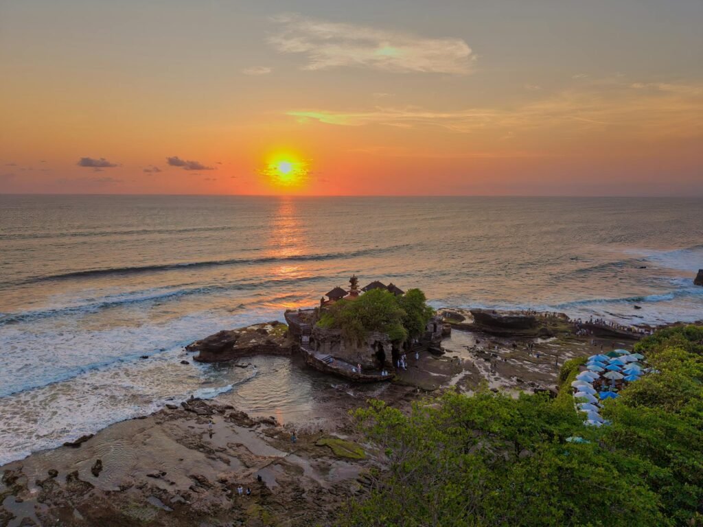 Sunset over Bali's rocky coastline with ancient temple structure surrounded by ocean waves and blue umbrellas.