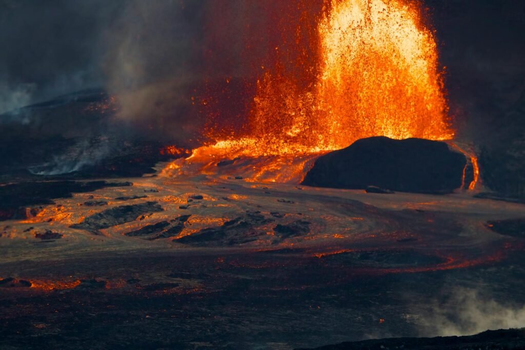 Dramatic lava fountain erupting from volcanic crater with glowing molten lava flows across dark ground.