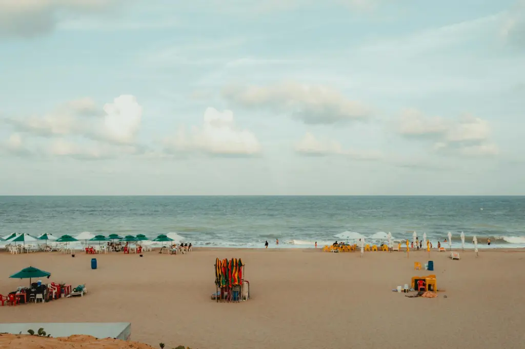 Wide sandy beach with colorful umbrellas, playground equipment, and swimmers enjoying the ocean under a partly cloudy sky.