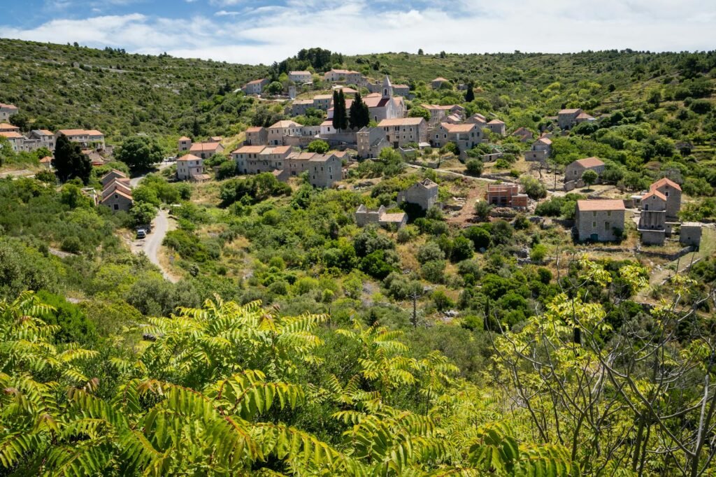 Mediterranean village nestled in lush green hills with stone houses and a church with tall spires.