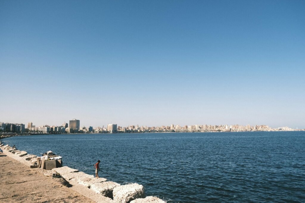 Person standing on rocky beach with Alexandria skyline and Mediterranean Sea under clear blue sky.