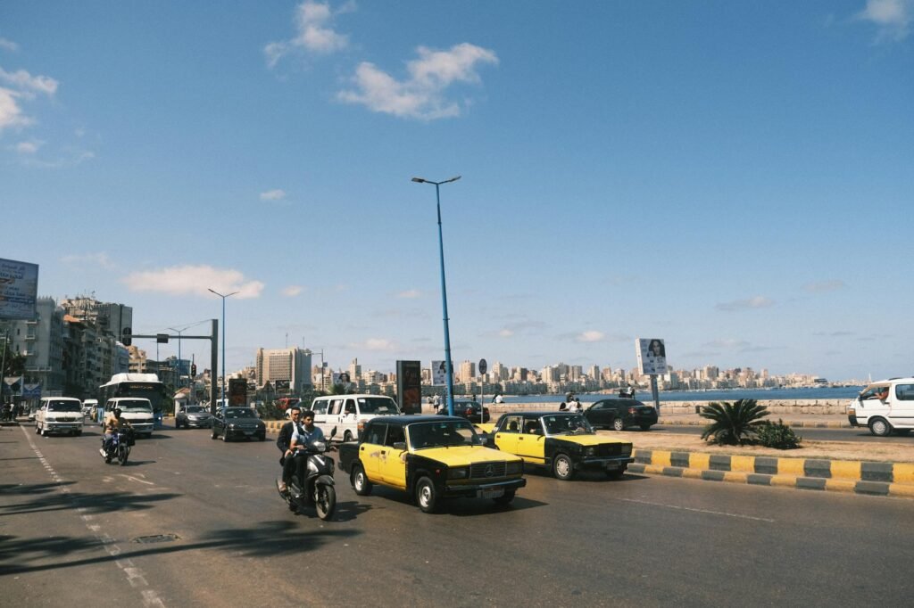 Busy street scene in Alexandria with yellow and black taxis, motorcycles, and city skyline visible across the water.