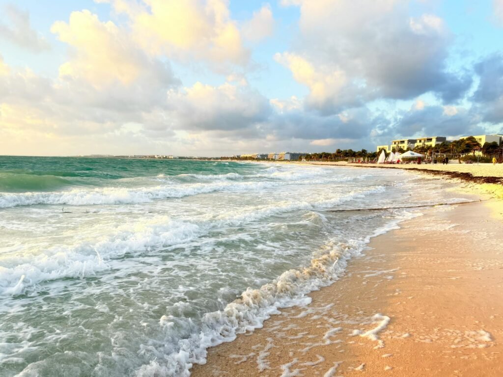Turquoise ocean waves rolling onto sandy beach with coastal buildings and cloudy sky in background.