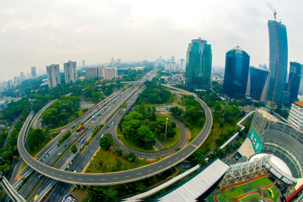 Aerial view of Jakarta cityscape with elevated highways, green park, modern skyscrapers, and distant skyline under cloudy…
