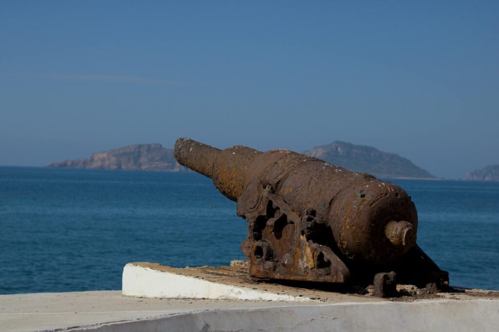 Rusted cannon relic on beach sand with blue ocean and distant islands in background.