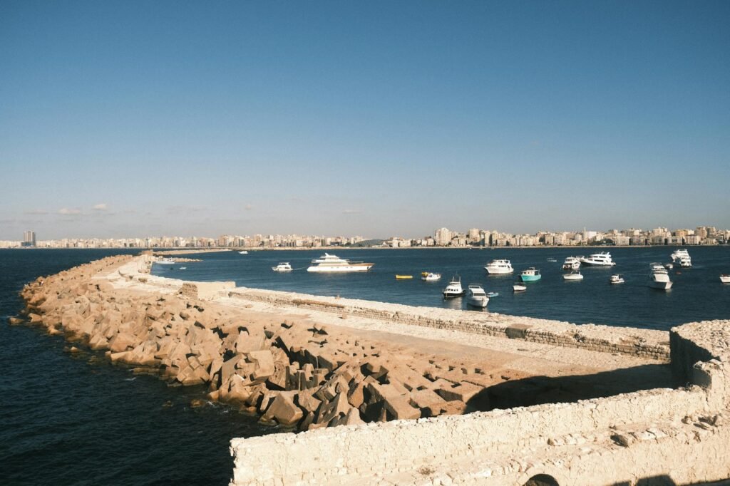 Rocky breakwater with moored boats in Alexandria's harbor and cityscape under clear blue sky.