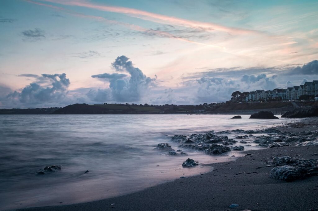 Coastal beach at sunset with calm waters, rocky shoreline, and pastel clouds over distant houses.